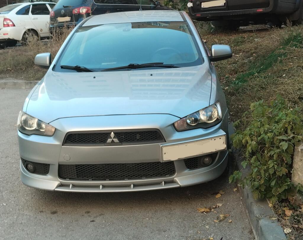 Серебристый автомобиль Mitsubishi Lancer, припаркованный у бордюра рядом с зарослями