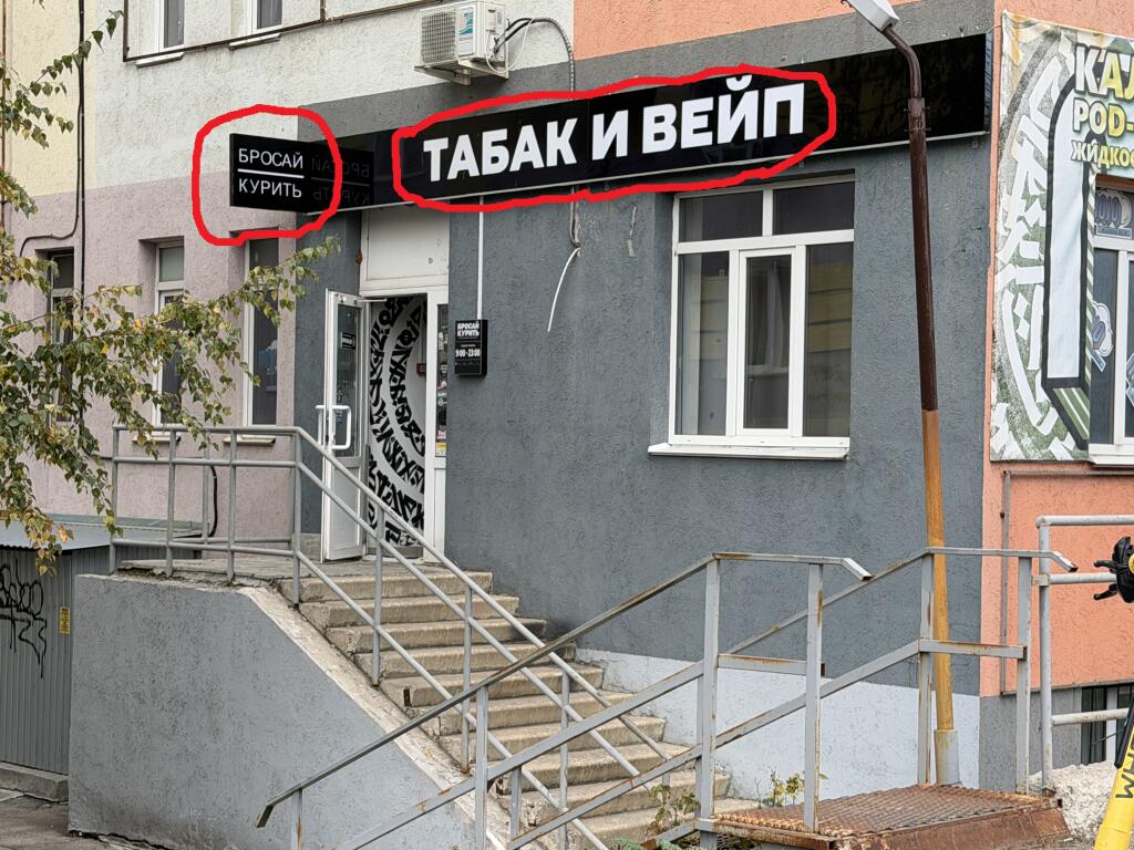 Фасад серого здания с вывесками «Бросай курить» и «Табак и вейп», обведенными красным маркером