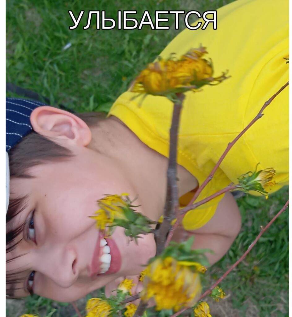 Мальчик в желтой футболке и бандане улыбается, наклонившись к цветущим растениям на траве, сверху надпись «улыбается